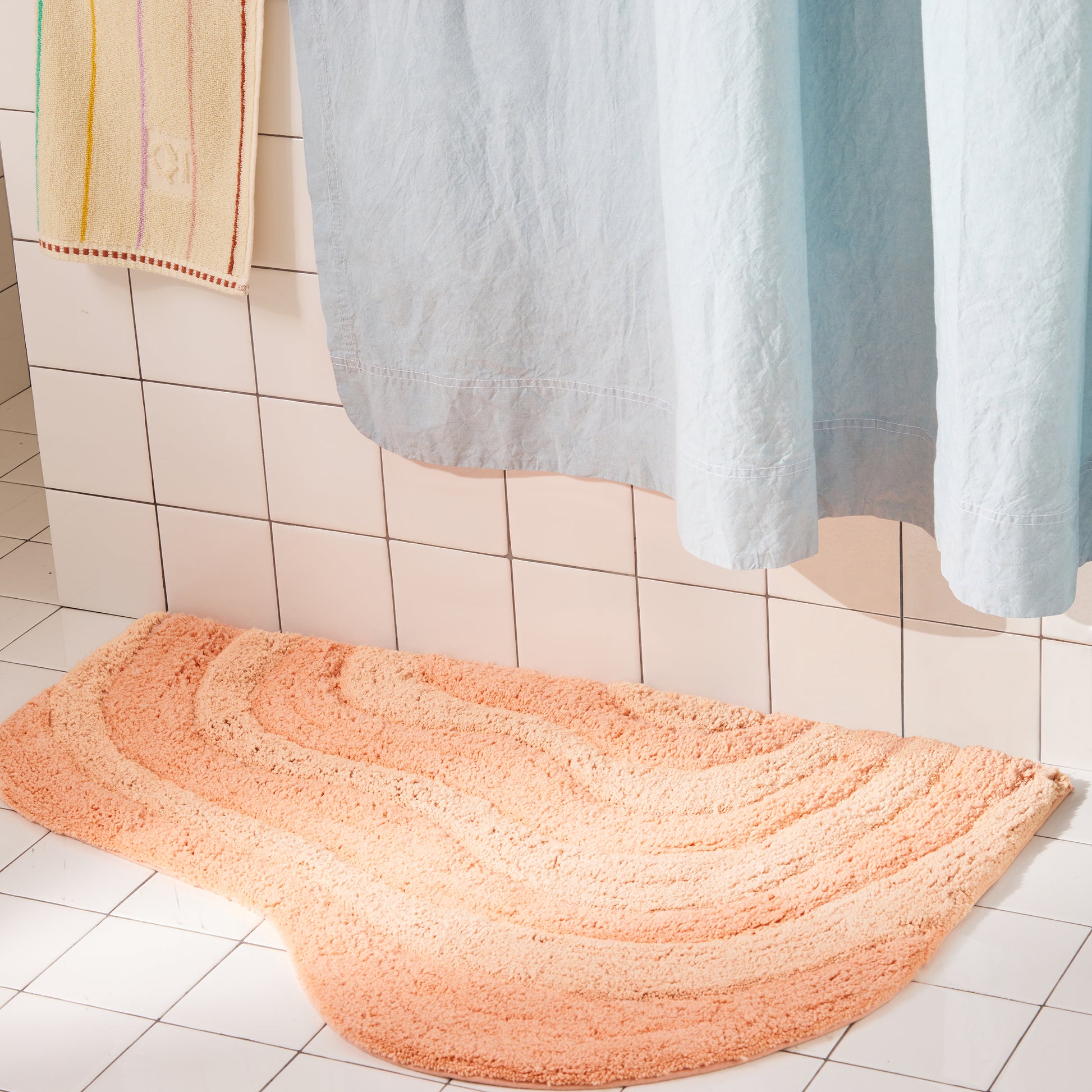 Peach curvy bath mat on a tiled bathroom floor with a shower curtain in the background.