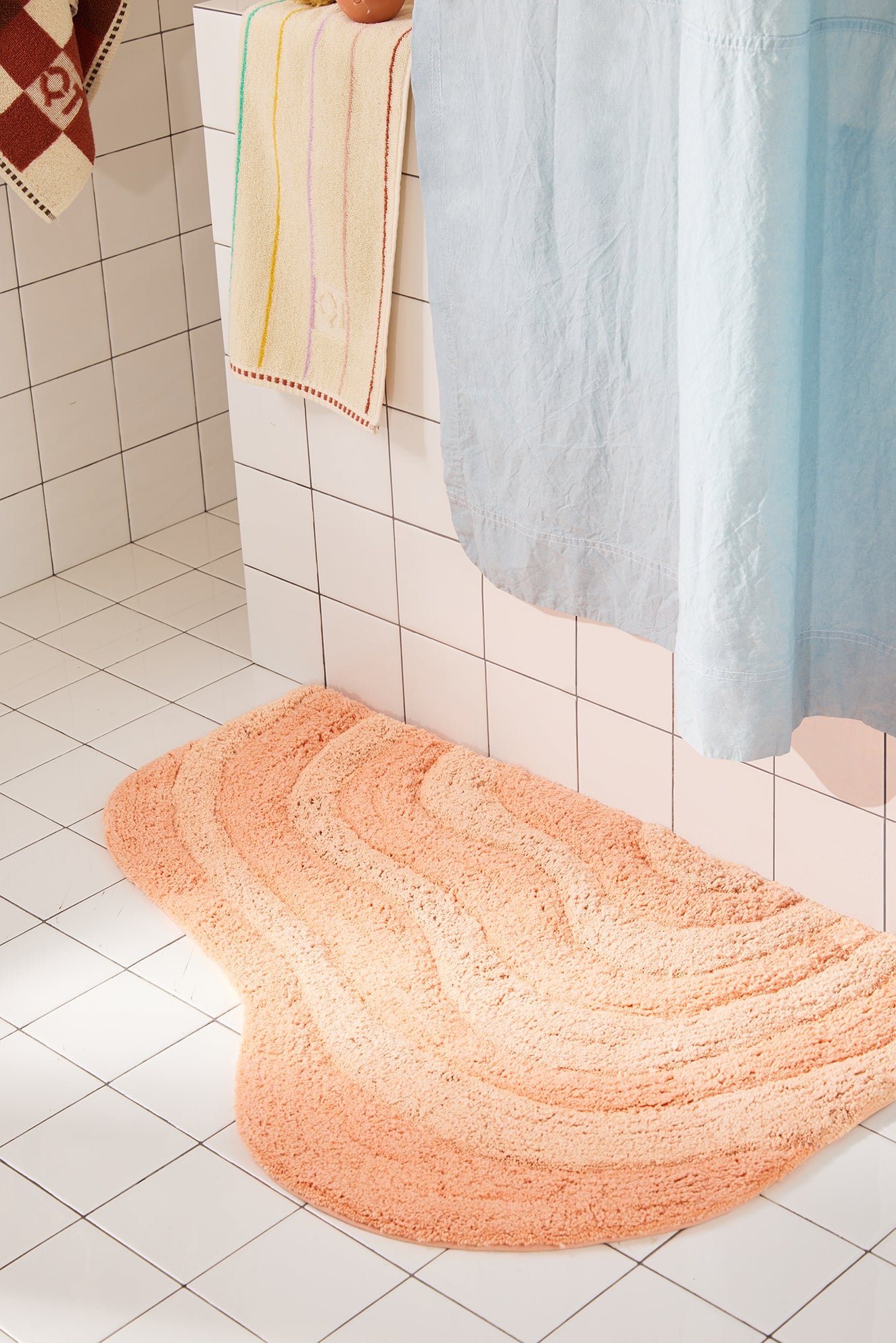 Peach Bath mat on a tiled bathroom floor with towels and shower curtain in the background
