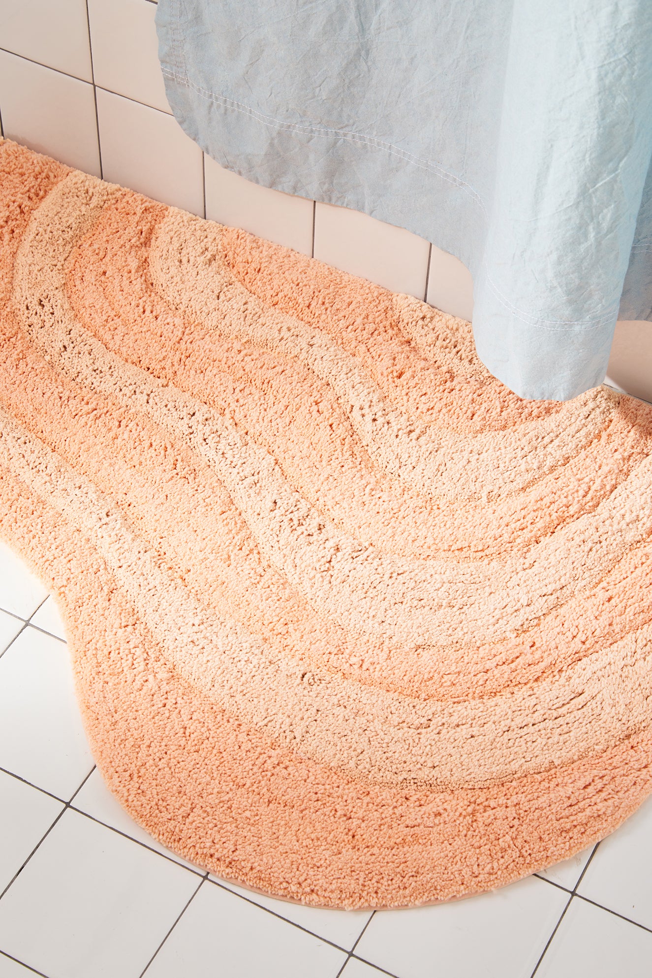 Peach bath mat on a tiled bathroom floor with a shower curtain in the background.