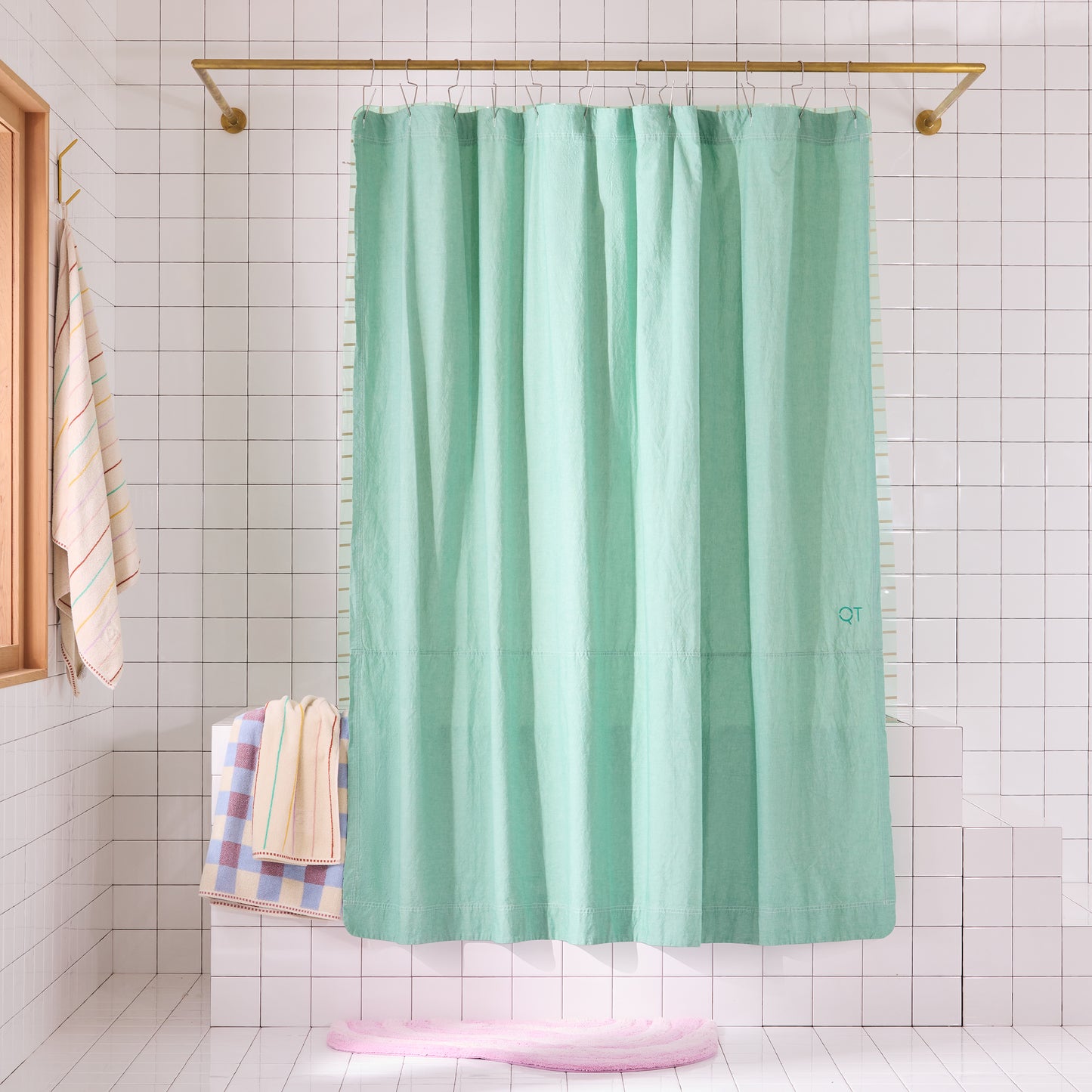 Green shower curtain hanging on a gold rod in a bathroom with tiled walls and a window. A pink rug and 