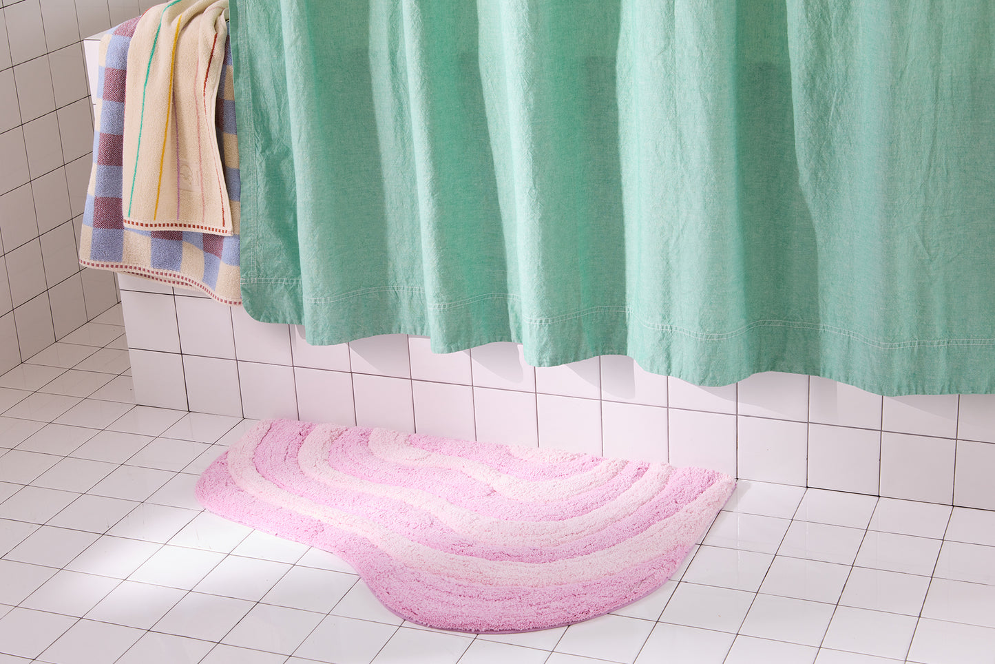 Pink bath mat on a tiled bathroom floor with a green shower curtain and colorful towel.