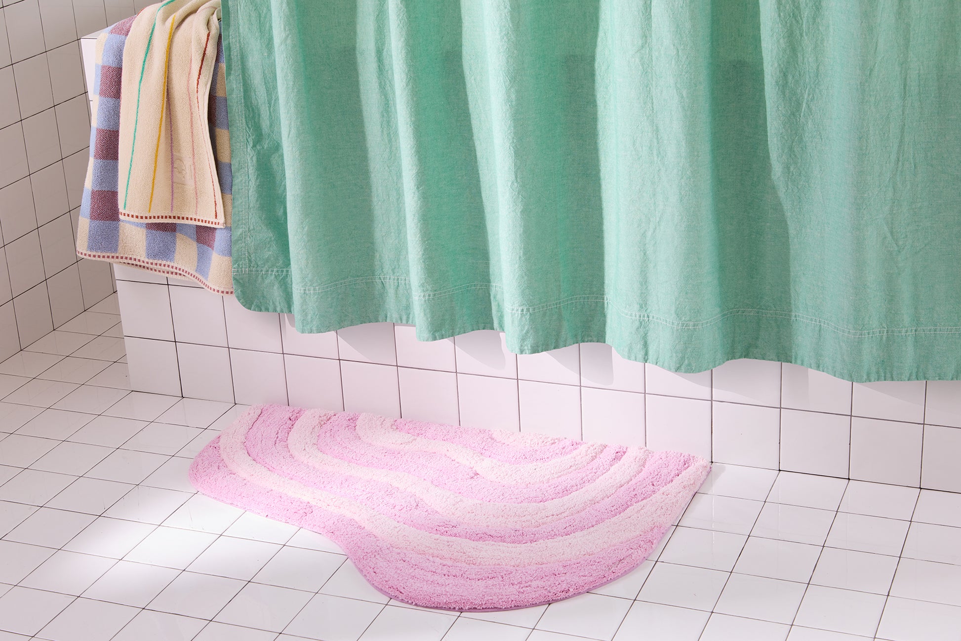 Pink bath mat on a tiled bathroom floor with a green shower curtain and colorful towel.