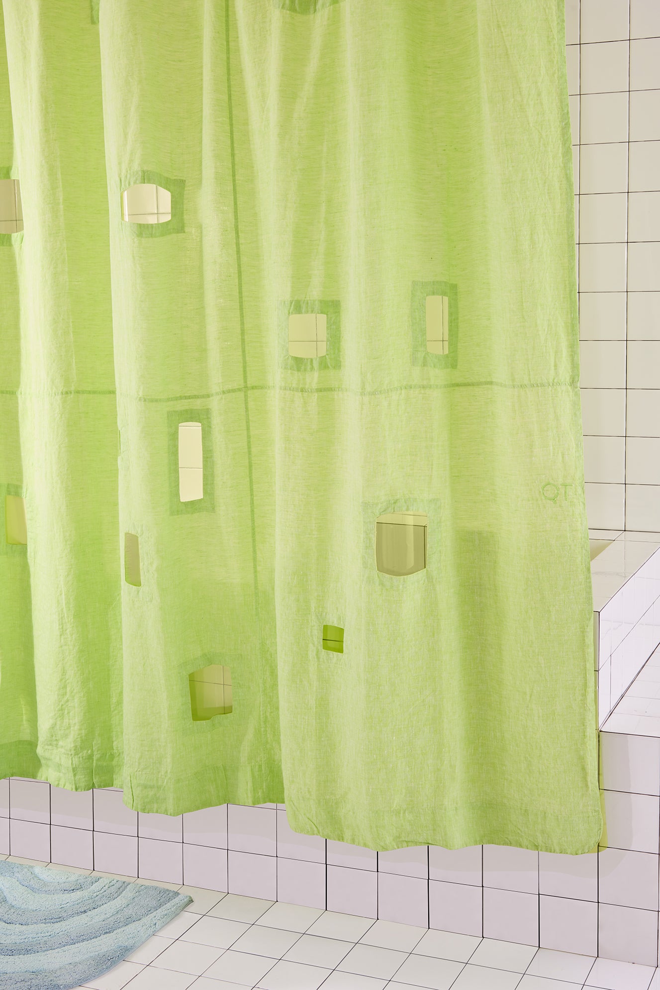 green shower curtain with cut outs and blue rug in white tiled bathroom. 