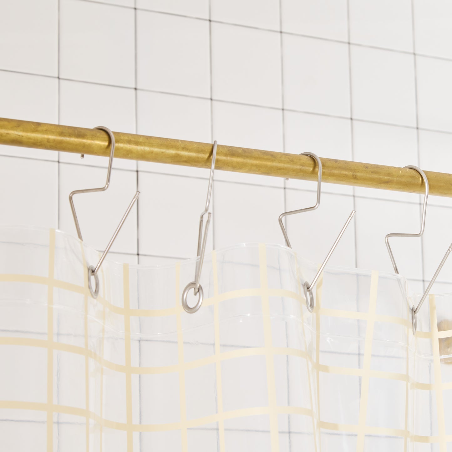 clear shower curtain with cream grid pattern on Metal hooks on a brass rod against a tiled wall background