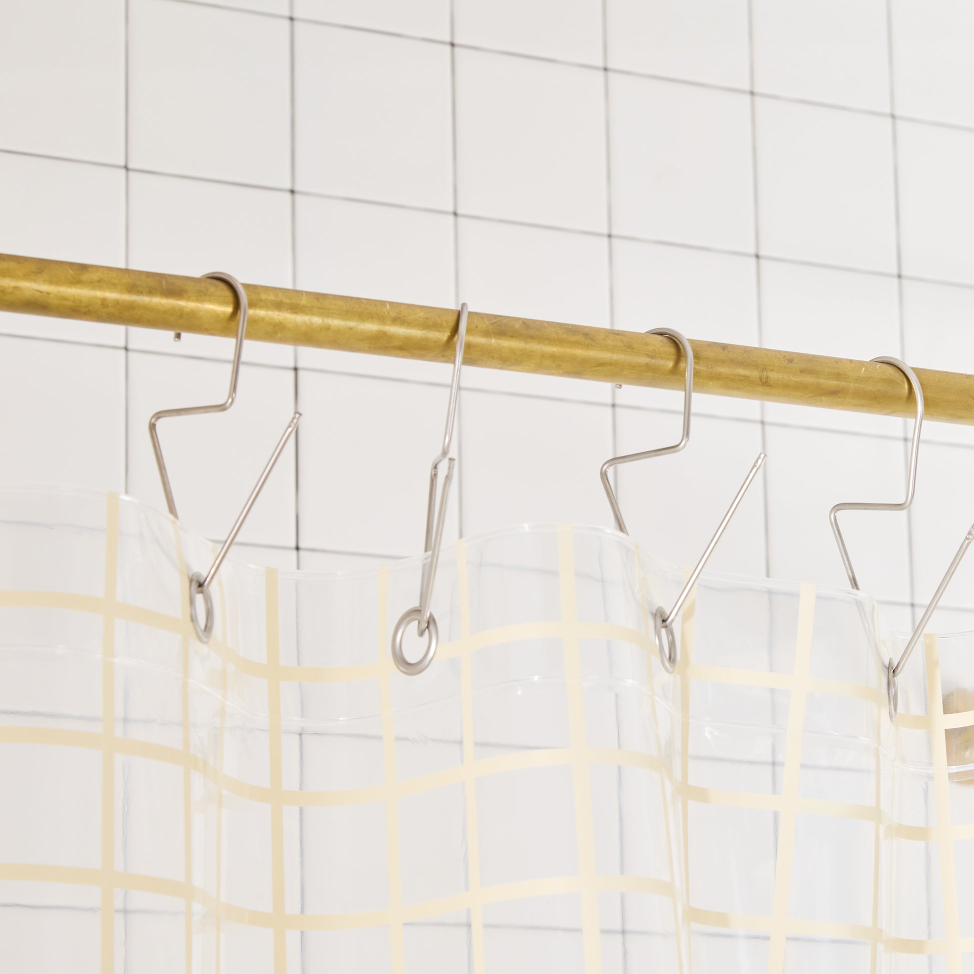 clear shower curtain with cream grid pattern on Metal hooks on a brass rod against a tiled wall background