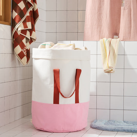 pink and cream laundry bin with burgundy nylon straps in white tiled bathroom. 