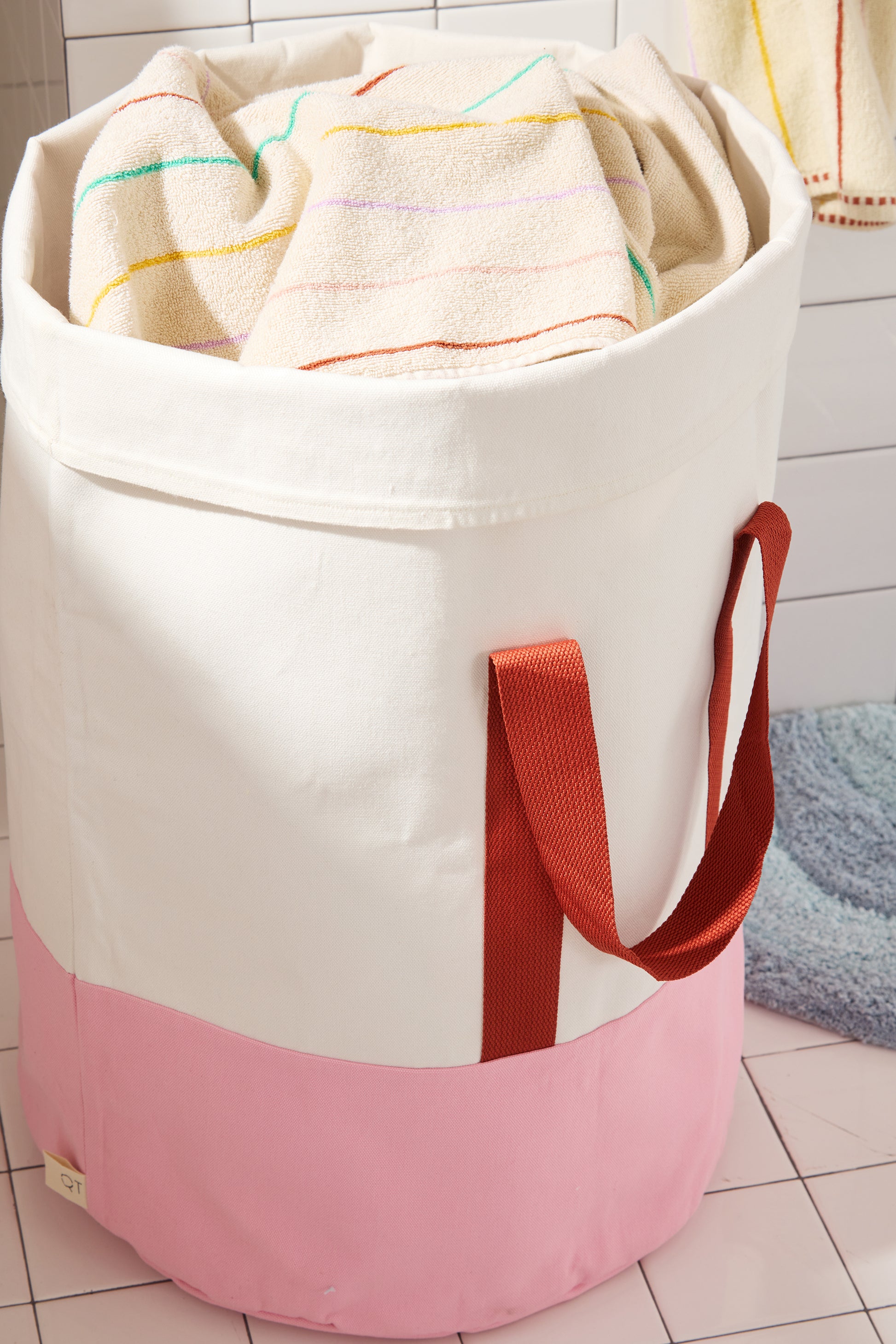 pink and cream laundry bin with burgundy nylon straps in white tiled bathroom. 