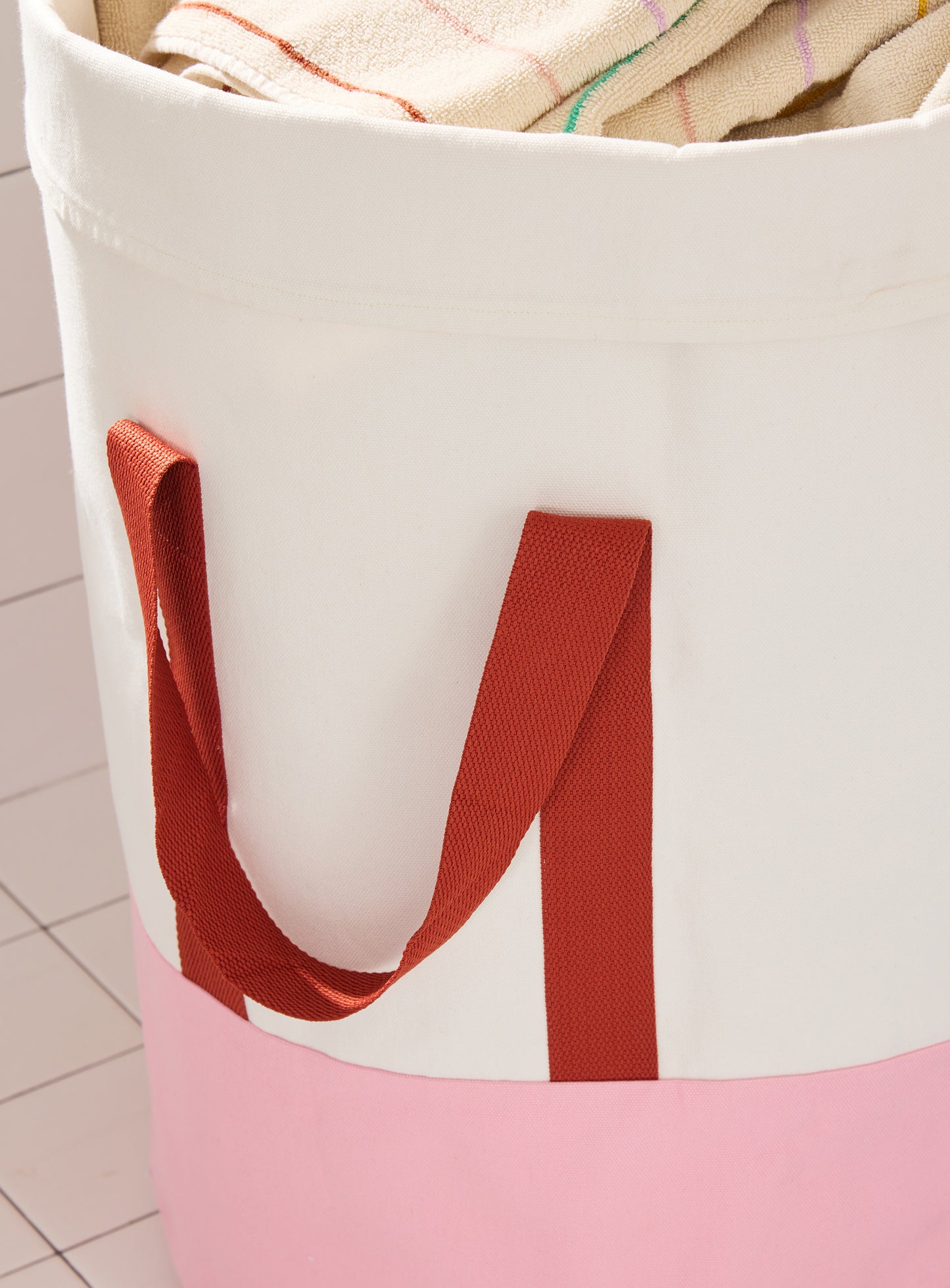 pink and cream laundry bin with burgundy nylon straps in white tiled bathroom and striped towel. 