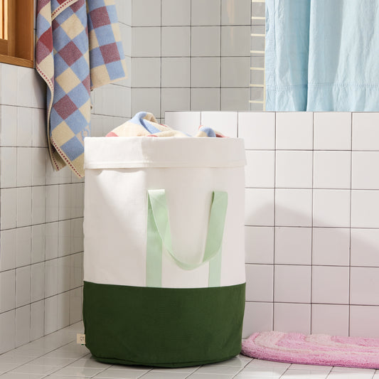 Green and cream color blocked laundry hamper in white tiled bathroom. 