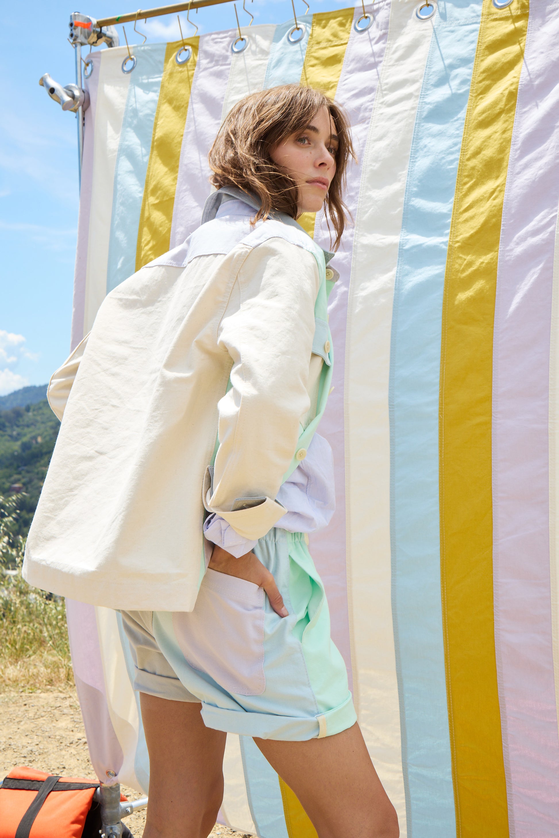 woman against a striped background wearing a cream jacket adn colorful shorts. 