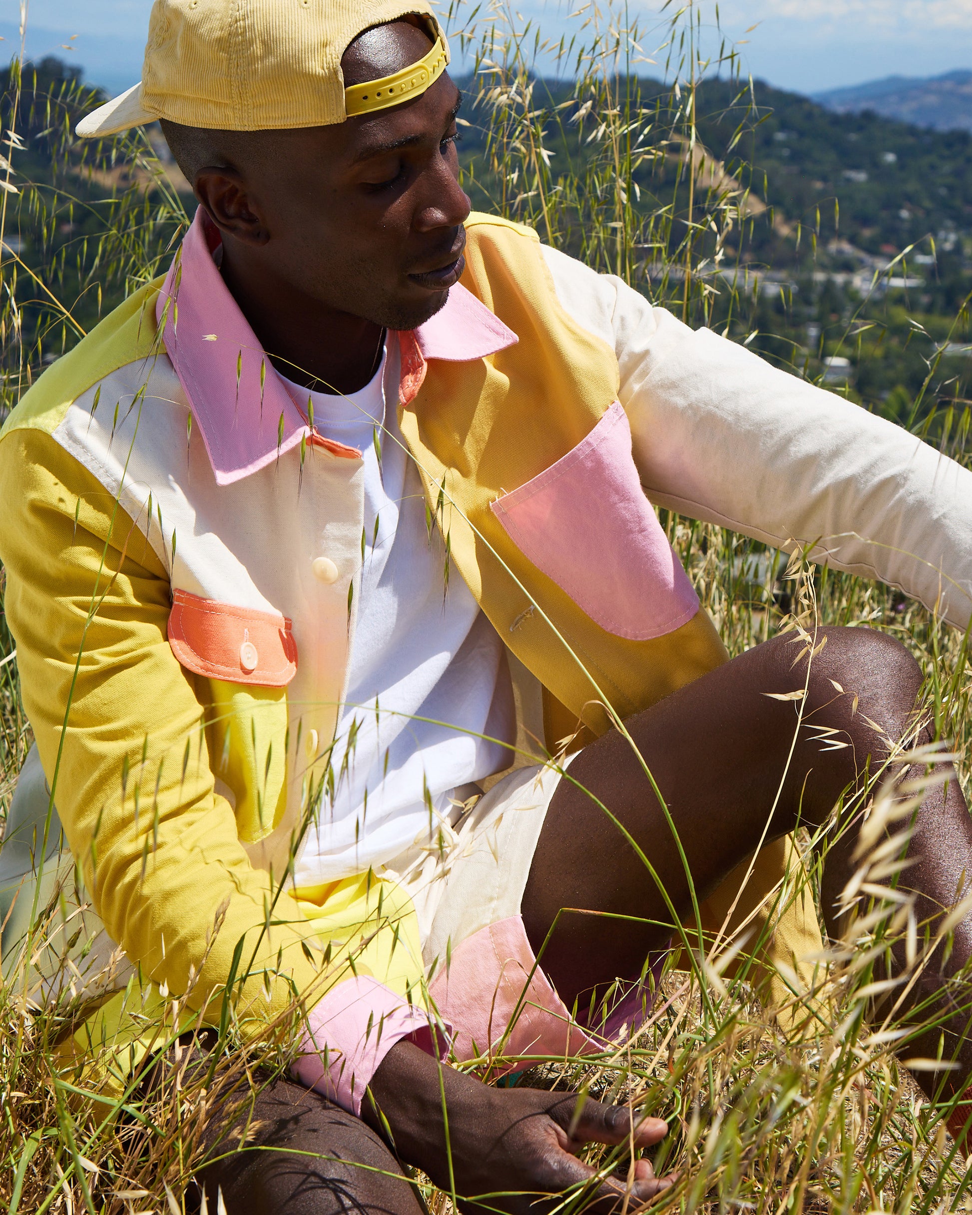 Person wearing a colorful jacket and yellow cap sitting in tall grass with a scenic background