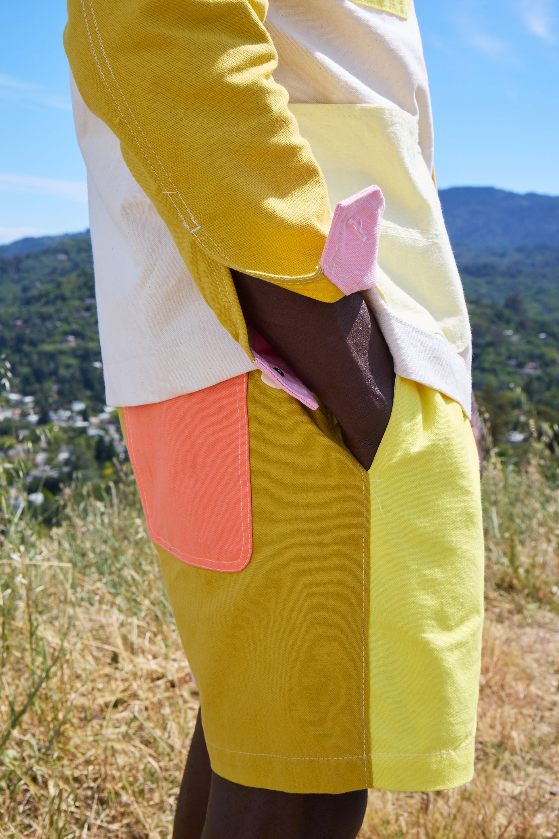 Person wearing a colorful jacket with a pink patch on a yellow tow-toned short, standing on a hill with mountains in the background.