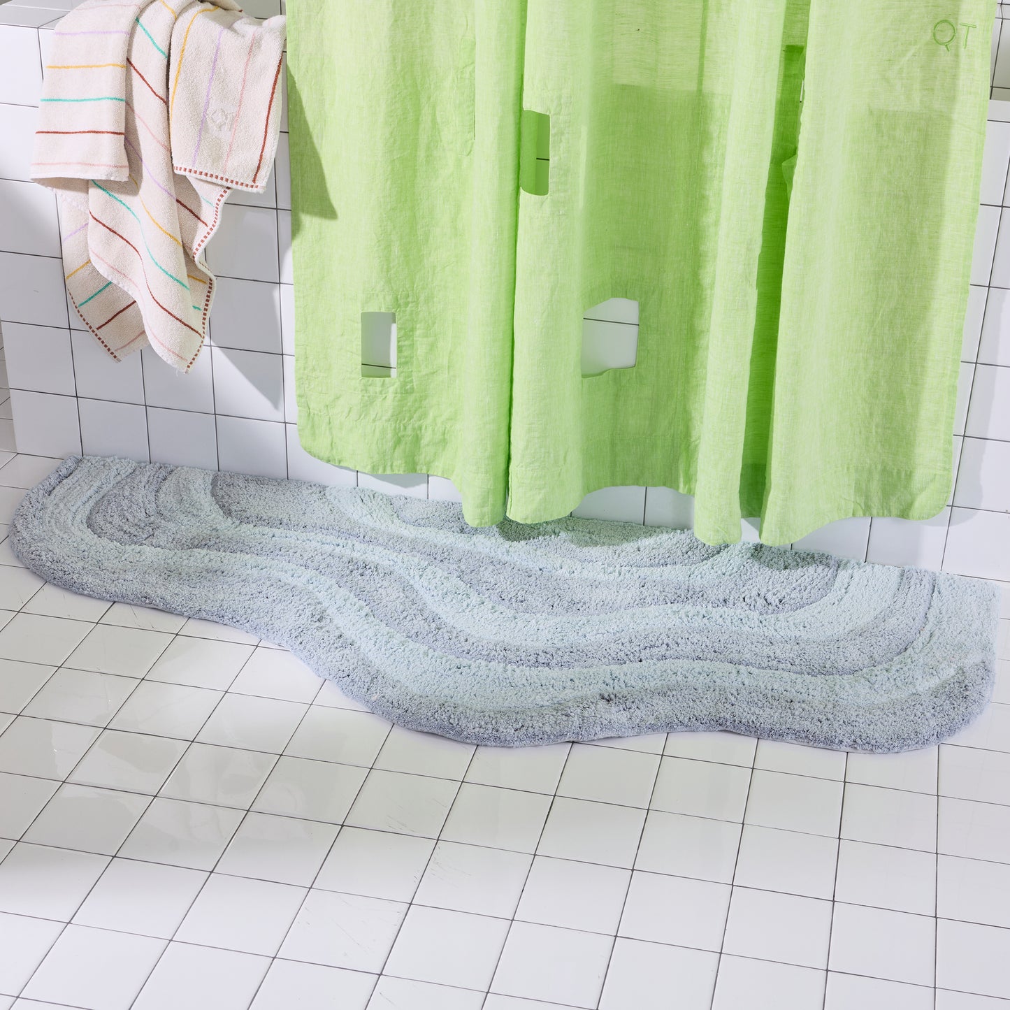 Green shower curtain with a light blue wavy bath mat on a tiled bathroom floor.