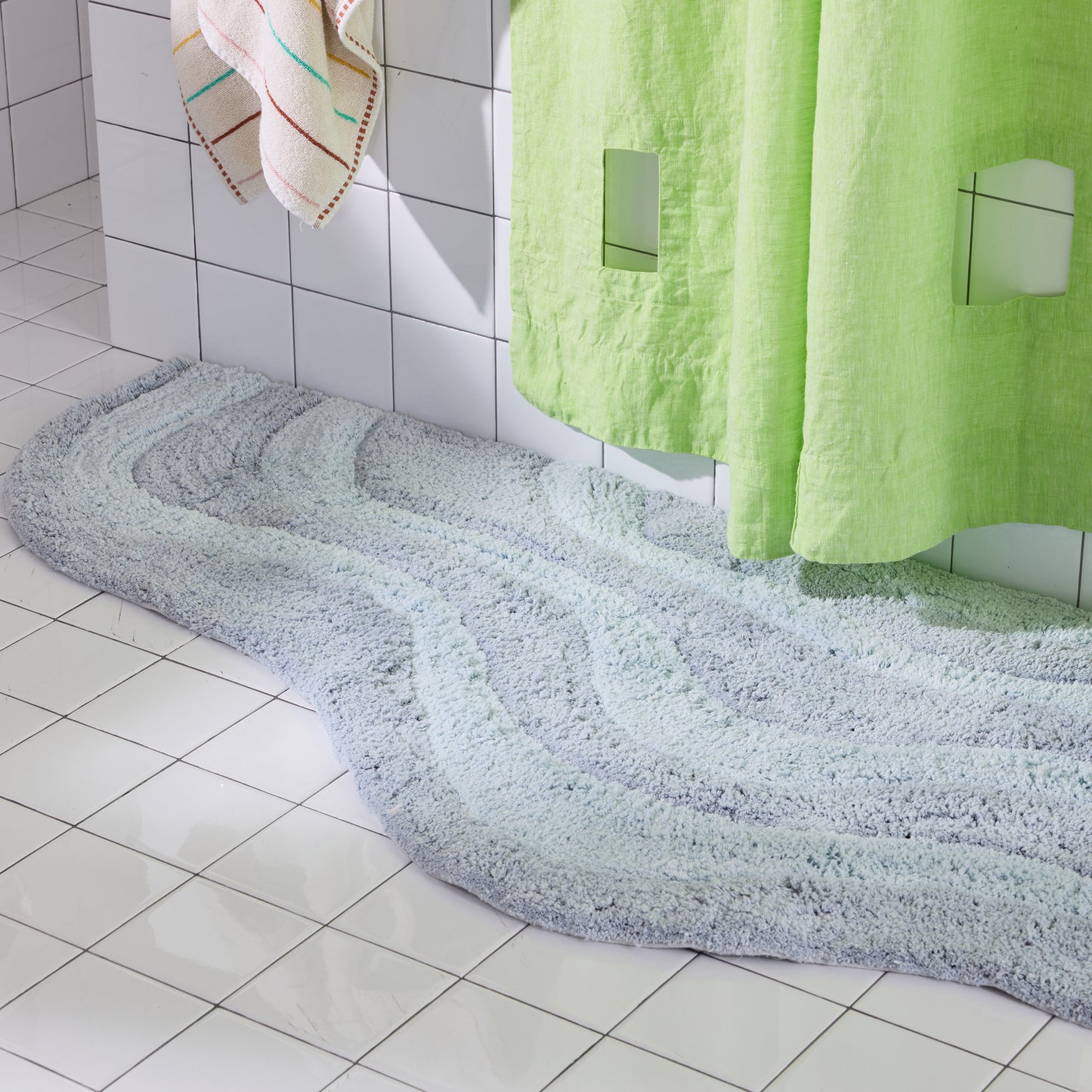 Light blue Bath mat on a tiled bathroom floor with green shower curtain and towels.