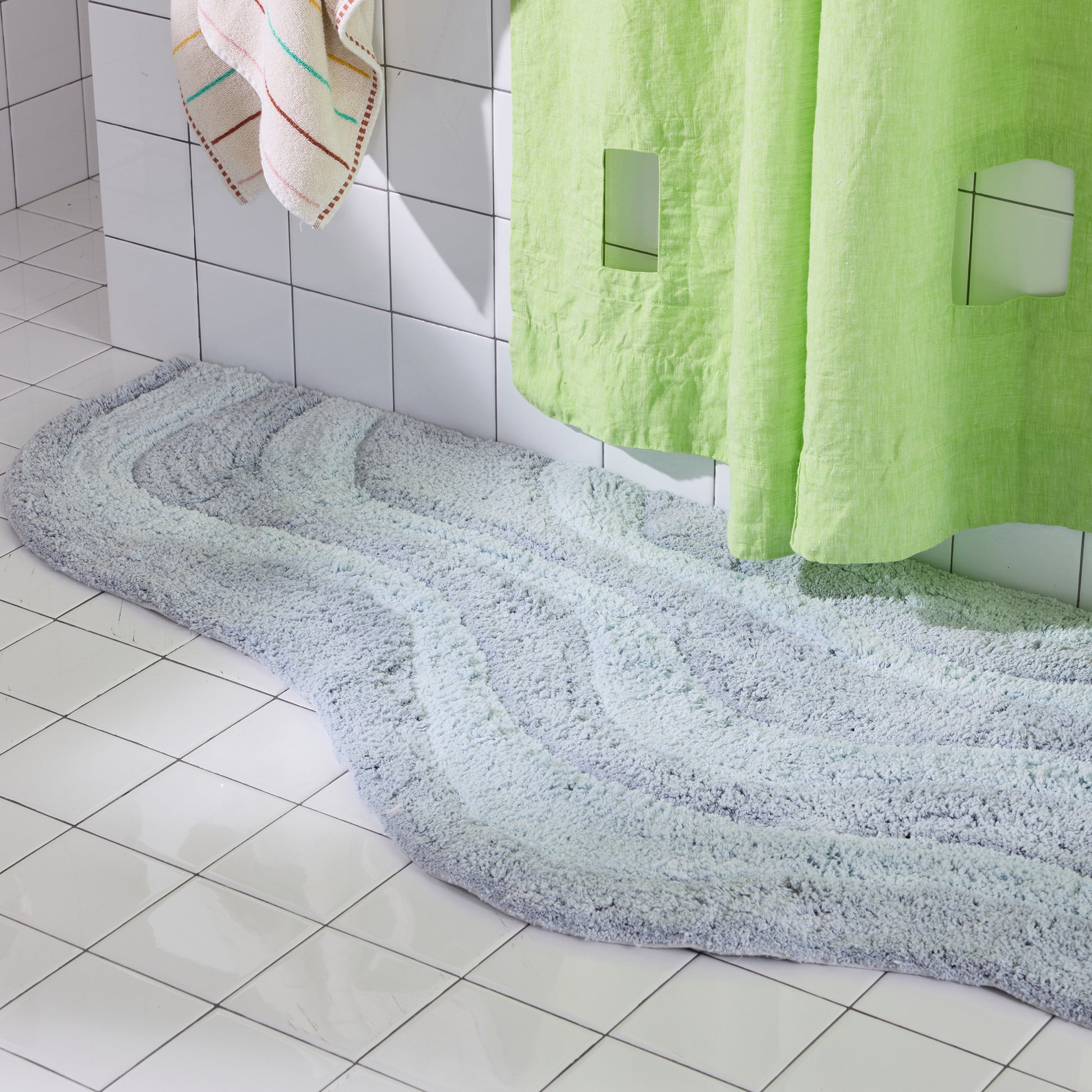 Light blue Bath mat on a tiled bathroom floor with green shower curtain and towels.