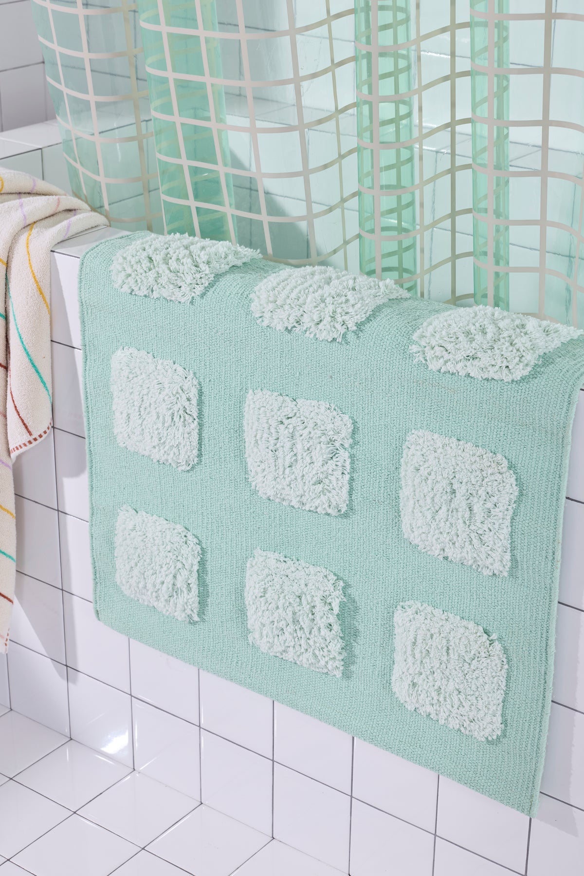 Close up of Mint/Teal bath rug with raised tufted squares in natural tones, draped over a white tiled bathroom ledge beneath a teal shower curtain.