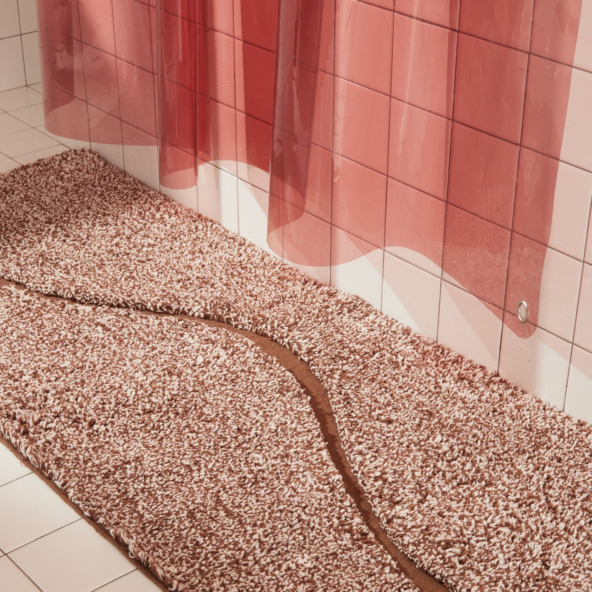 Brown bath mat in 56"x22" size with curvy path stripe contrast shot styled in a white tiled bathroom next to rose colored sun shower curtain.