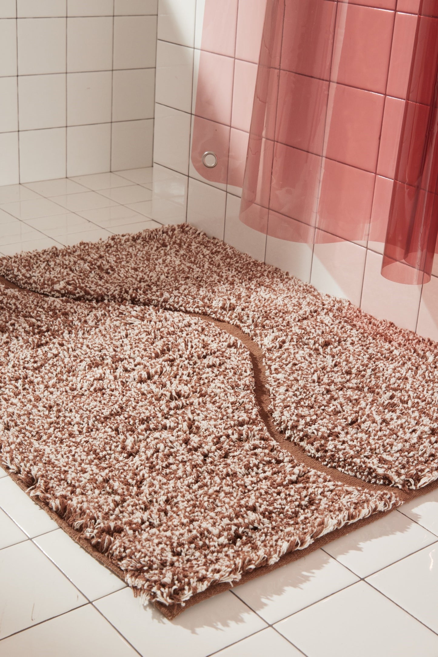 Side view of brown bath mat in 21"x31" size with curvy path stripe contrast shot styled in a white tiled bathroom next to rose colored sun shower curtain.