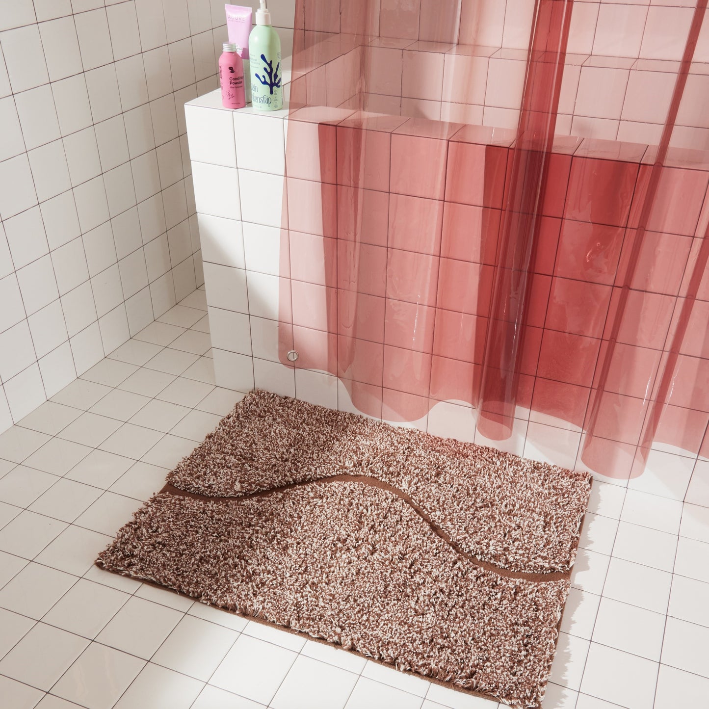 Brown bath mat in 21"x31" size with curvy path stripe contrast shot styled in a white tiled bathroom next to rose colored sun shower curtain.